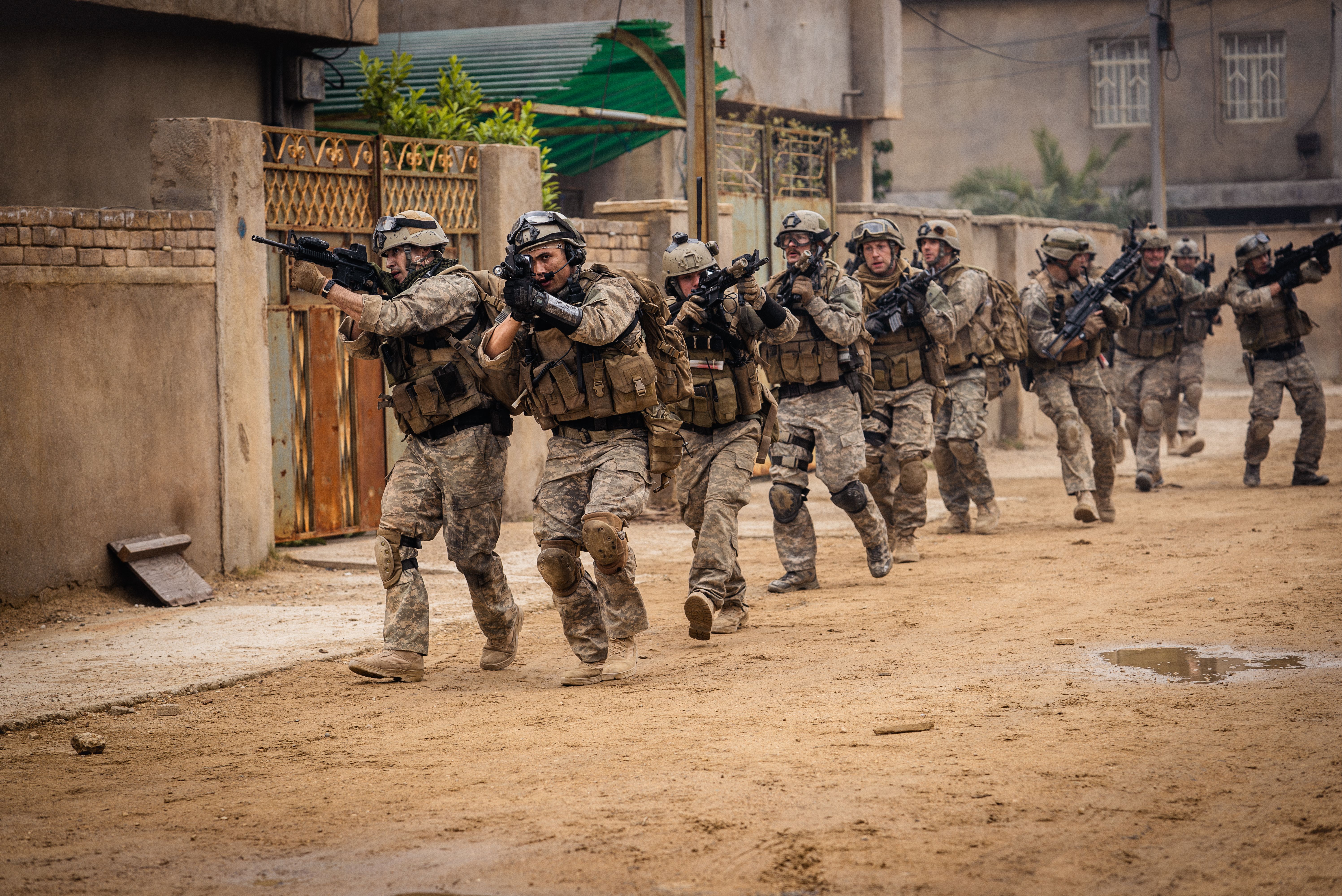 A team of soldiers in full gear in a linear formation moving along a street.