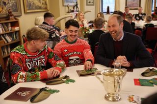 Prince William sitting at a table with two men wearing Christmas sweaters and laughing