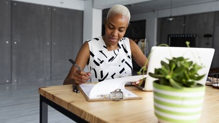 A woman sits at her dining room table with her laptop open nearby as she goes over a checklist on a legal pad.