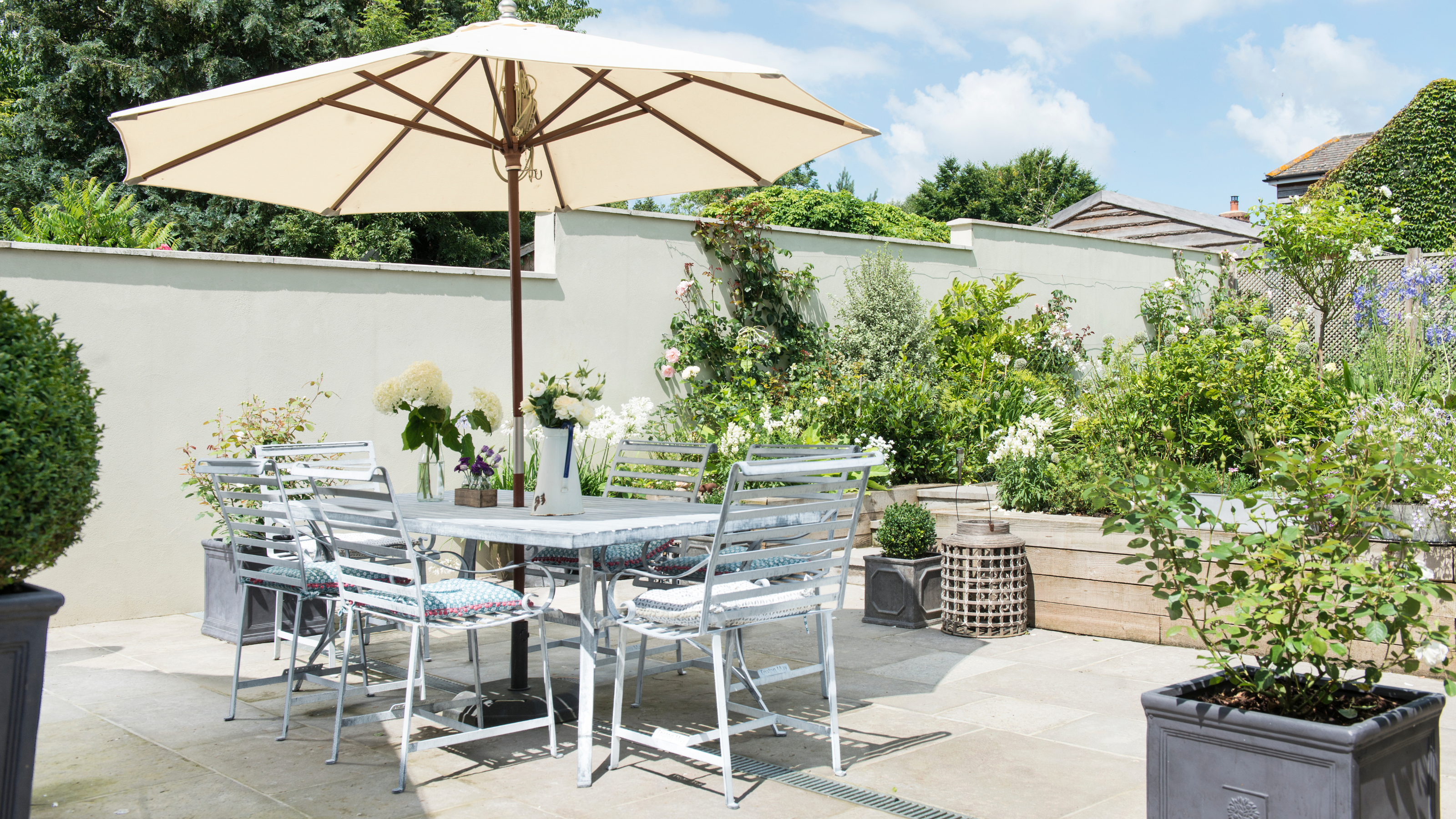Garden dining table and chairs complete with white parasol on a garden patio, surrounded by greenery in pots and planters.