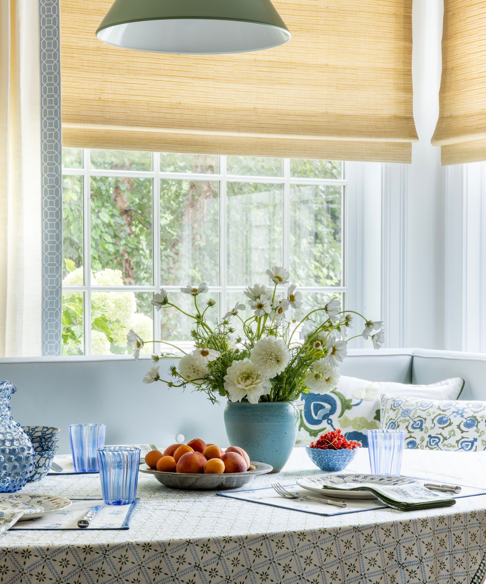 A high-angle shot of a breakfast nook. A light blue tufted banquette is tucked into a corner with large windows covered by woven straw-colored Roman shades. The table features a blue-and-white patterned tablecloth, blue fluted glasses, and a bowl of stone fruit. A teal ceramic vase holds a large arrangement of white flowers. A green pendant light hangs partially in view at the top.