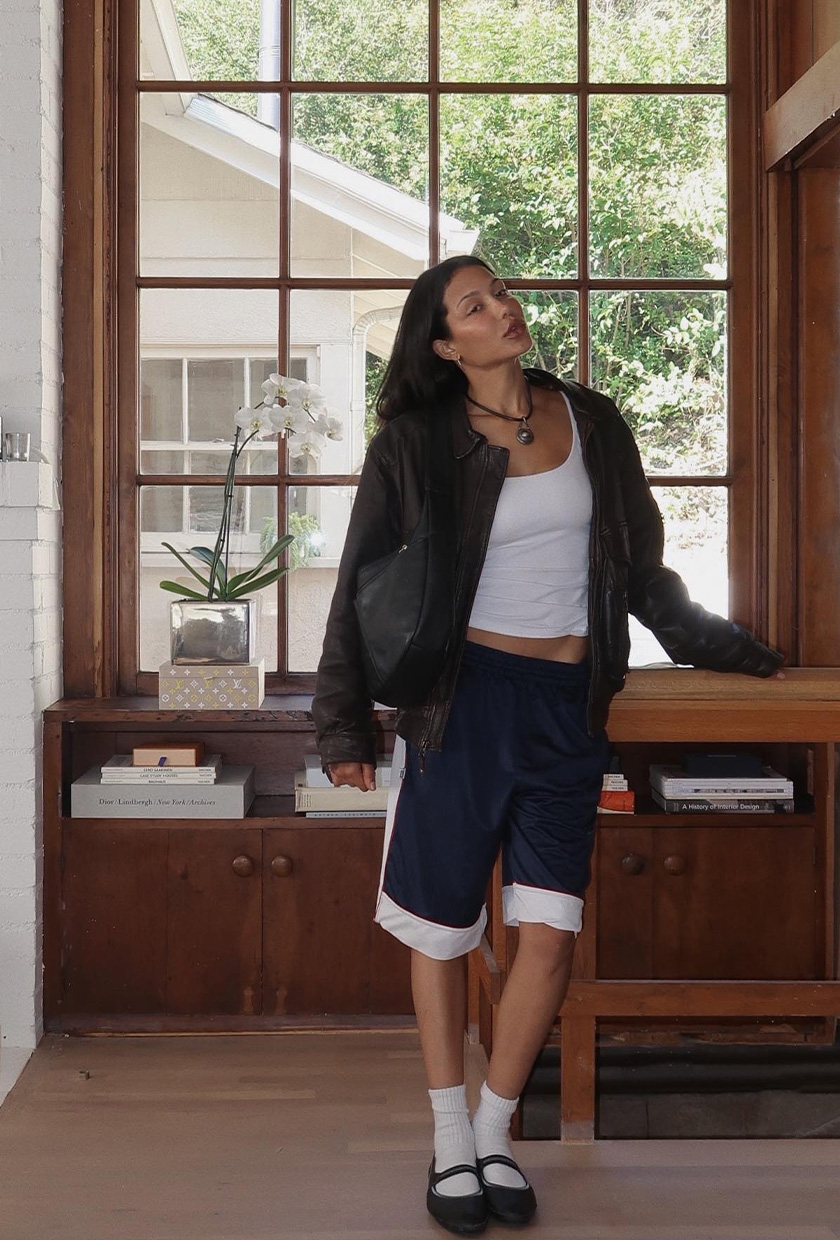 The best ballet sneakers are shown in a photo of a woman standing by a window inside the home wearing a leather jacket over a white tank top with basketball shorts styled with tall white socks, and mary jane sneakers