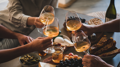 Glasses of orange wine toasting over a table with grapes and cheese.