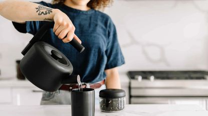 A female with curly hair wearing a navy blue t-shirt pouring hot water from a fellow clyde stovetop tea kettle