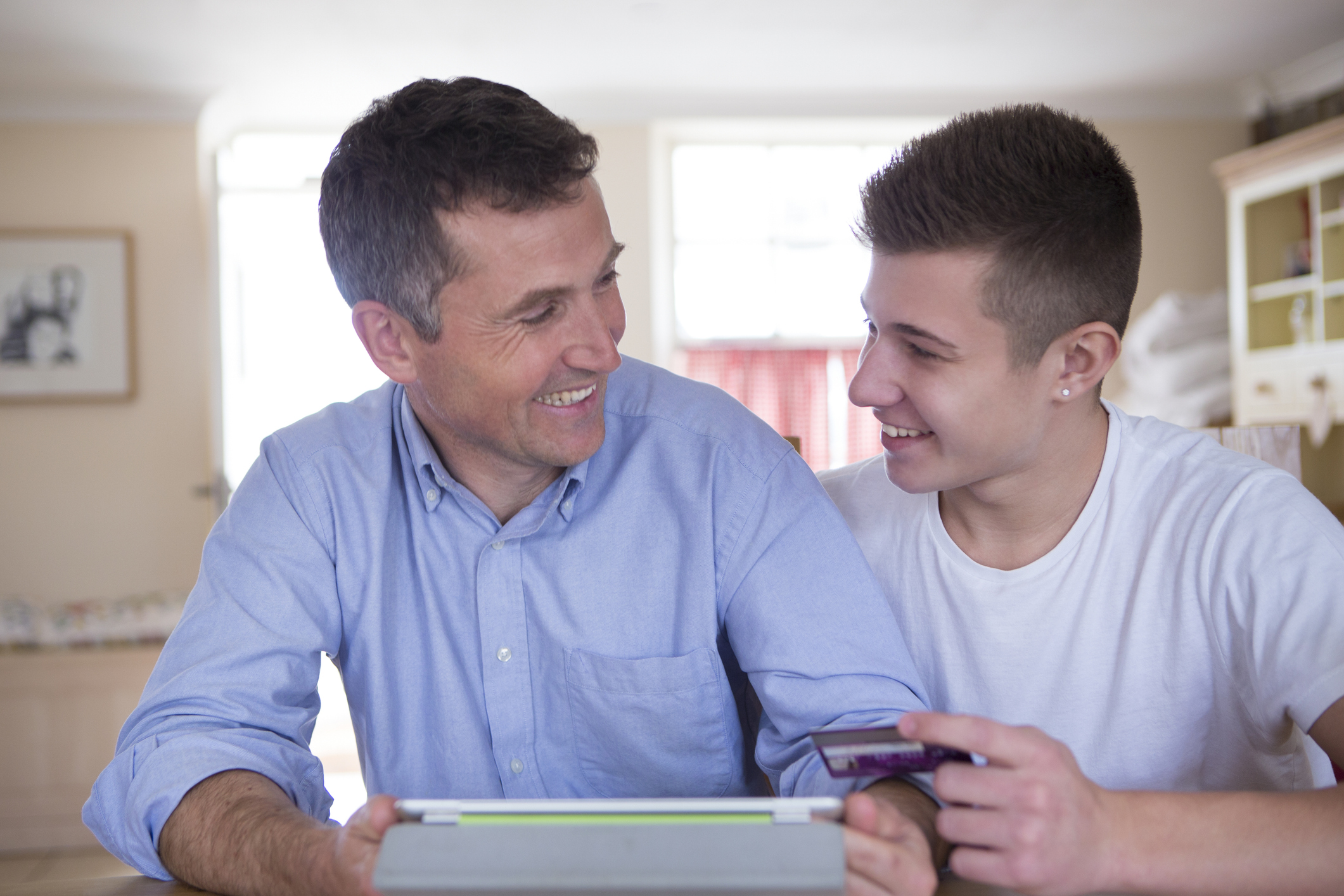 A middle aged father and teen son at home. They are looking at each other and smiling and they are holding a digital tablet.