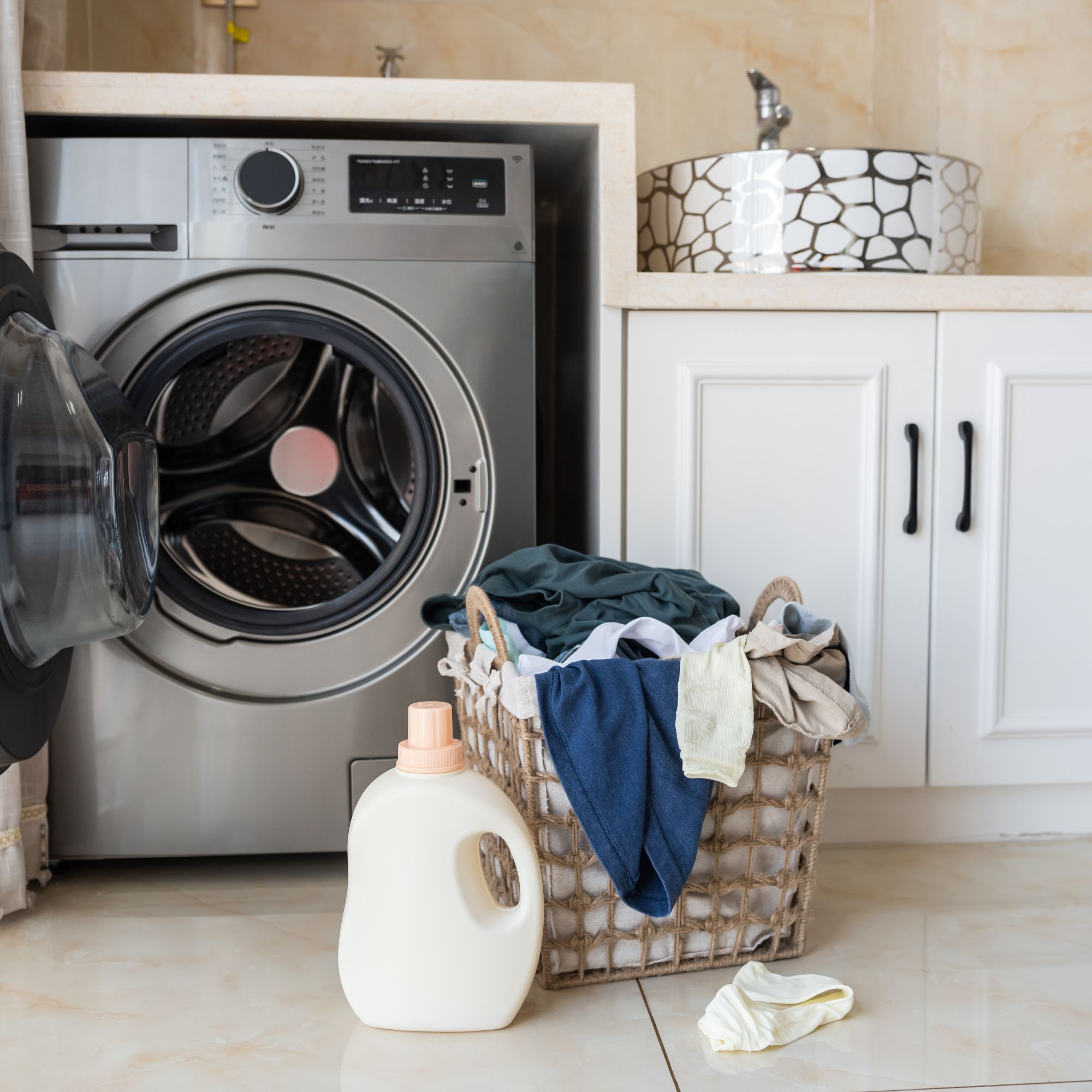Open grey washing machine in utility room with basket of laundry and bottle of detergent on the floor in front 