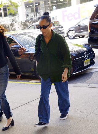 Zoe Kravitz wears black sunglasses, an oversize green button-down shirt, baggy blue jeans, and black slip-on vans.