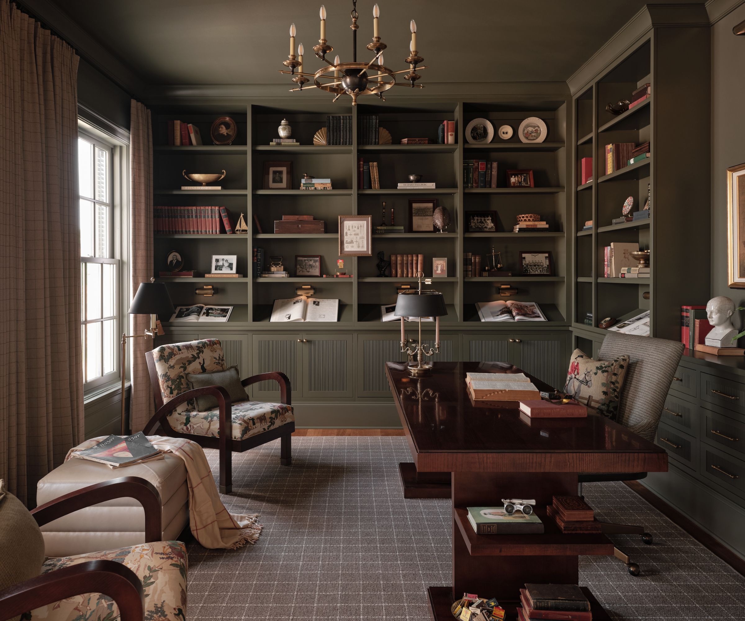 A home office with khaki color-drenched walls and built-ins, a neutral rug with a grid pattern, and a large wooden desk and two floral accent chairs.