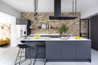 black kitchen units with white countertops, gray tiled floor, modern cooker hood, exposed brick wall, pendant lights, bar stools, kitchen island with hob and breakfast bar