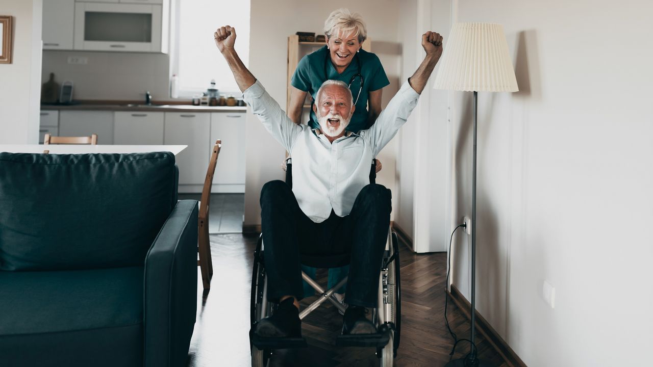 An older man in a wheelchair puts his hands in the air while a nurse pushes his chair in his apartment.