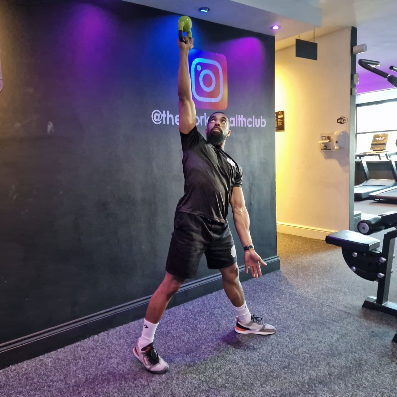 Renel Scarlett performs a kettlebell windmill. He is standing with his feet wider than shoulder-width apart. His left arm hangs at his side, while the right arm is held straight up overhead grasping a kettlebell. Behind him we can see the corners of some gym machinery.