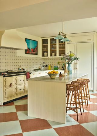 beadboard kitchen cabinets with chequered flooring and an island in the middle of the kitchen with bar stools placed next to it. Above the kitchen is a shaded pendant light.