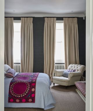 Coxy bedroom with dark gray painted walls, cream curtains, bed with white linen and pink throw, cream armchair