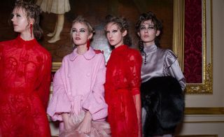 Four female models wearing looks from Simone Rocha's collection. Two models are wearing red semi-sheer dresses with sleeves and red detail. Another model is wearing a light pink belted coat with buttons. And the fourth model is wearing a grey high neck piece with sleeves and she is holding a black fur stole over her arm. All the models are wearing earrings
