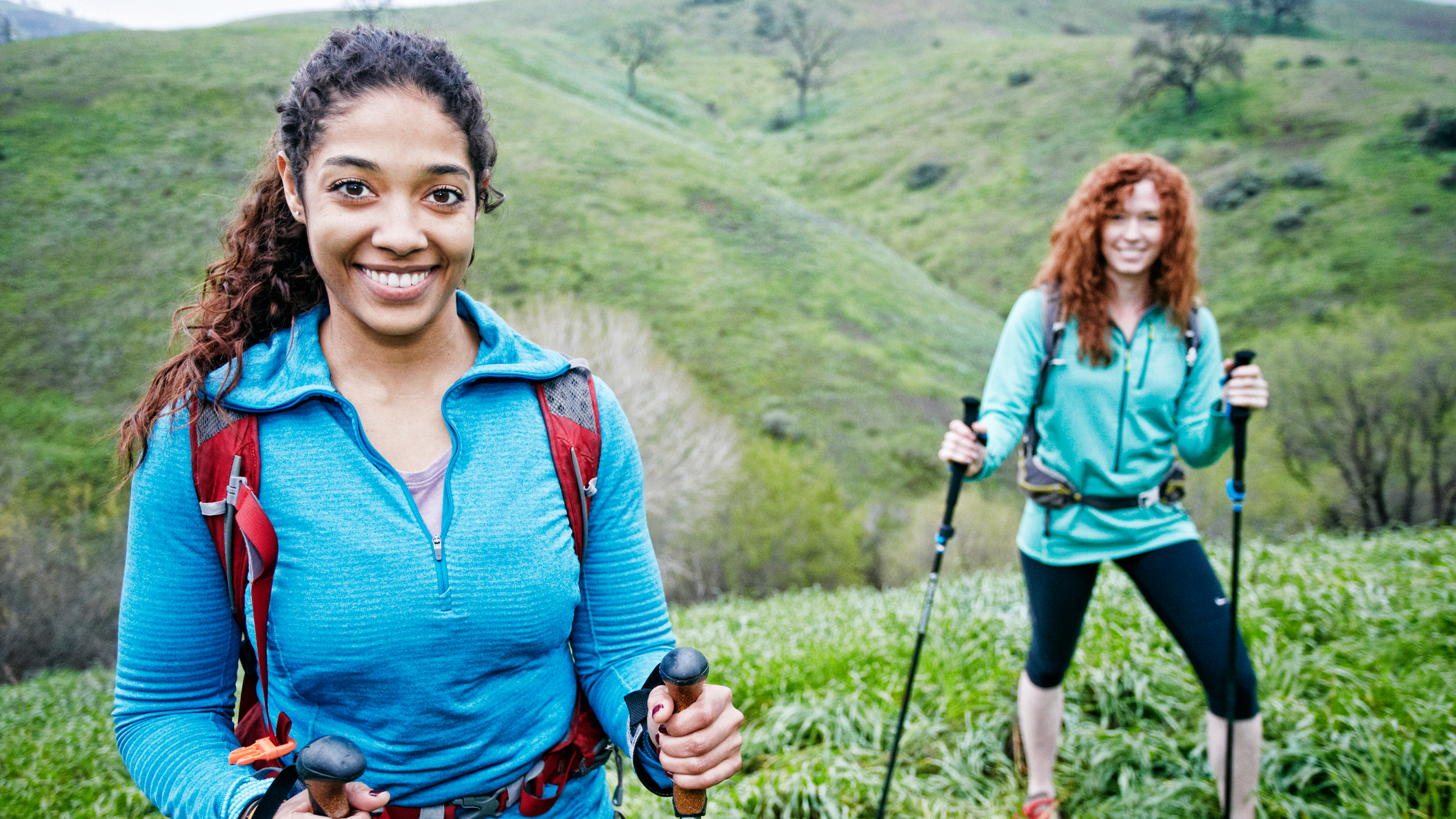 Two woman in hiking gear, including walking poles, smile at the viewer, with rolling green hills behind them
