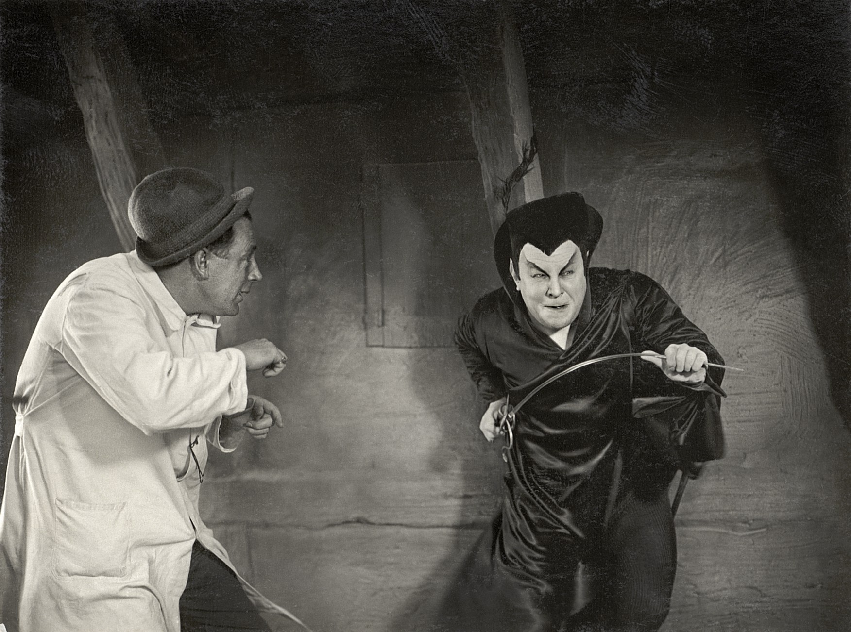 An actor in dark satin robes and dramatic make-up with a sharp widow&rsquo;s peak lunges forward holding a thin rapier, while a man in a white coat and hat gestures towards him on a stone-walled set.