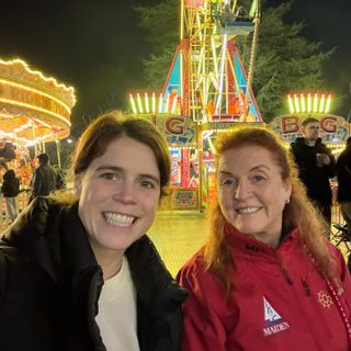 A selfie of Princess Eugenie and Sarah Ferguson with no makeup wearing winter coats and standing in front of lit-up fair rides at night