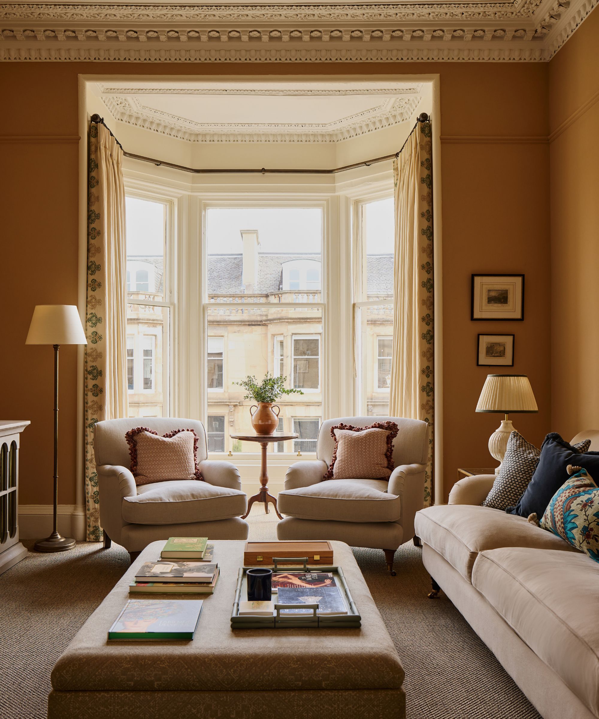 Warm ochre living room featuring a large bay window, two elegant beige armchairs, a central upholstered ottoman table, and traditional crown molding
