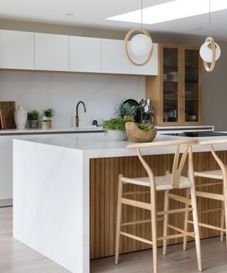 A white kitchen island with indoor plants and a bowl of limes on it with a wooden base, two curved Scandi-style seats behind it, two glass and wood circular pendant lights above it, and white cabinets behind it