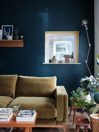 inky blue living room with sage green couch, shelf on wall, chrome wall light, wood coffee table, wooden stools, basket, coffee table books