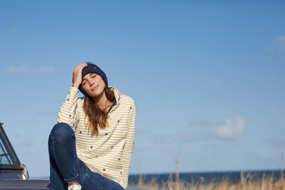 A woman sat in a field against a blue sky wearing a white and yellow striped top, blue jeans and a blue hat from Joules.