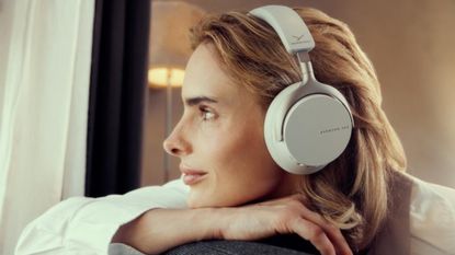 A woman with blonde hair leaning over the back of a sofa or chair while wearing a pair of Beyerdynamic Aventho 200 headphones