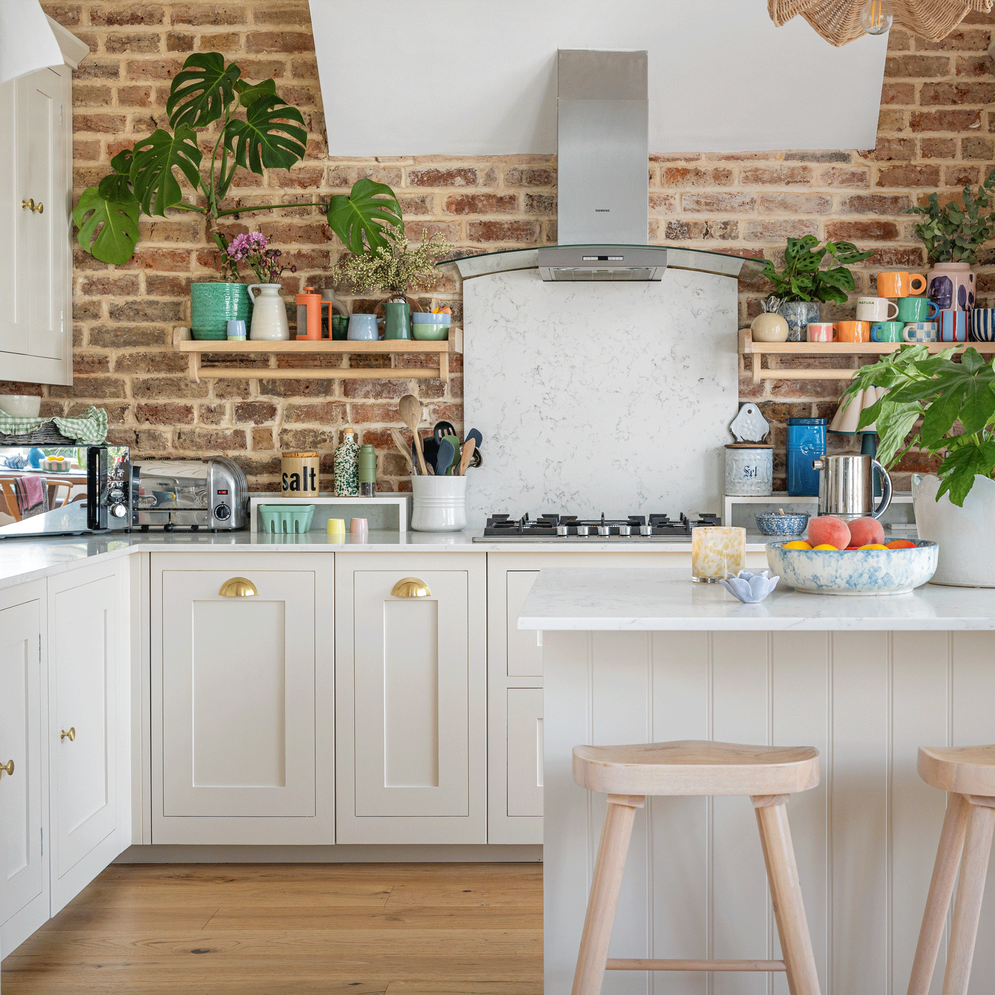 a neutral kitchen with cream coloured Shaker cabinetry with brass cup handles and an exposed brick wall