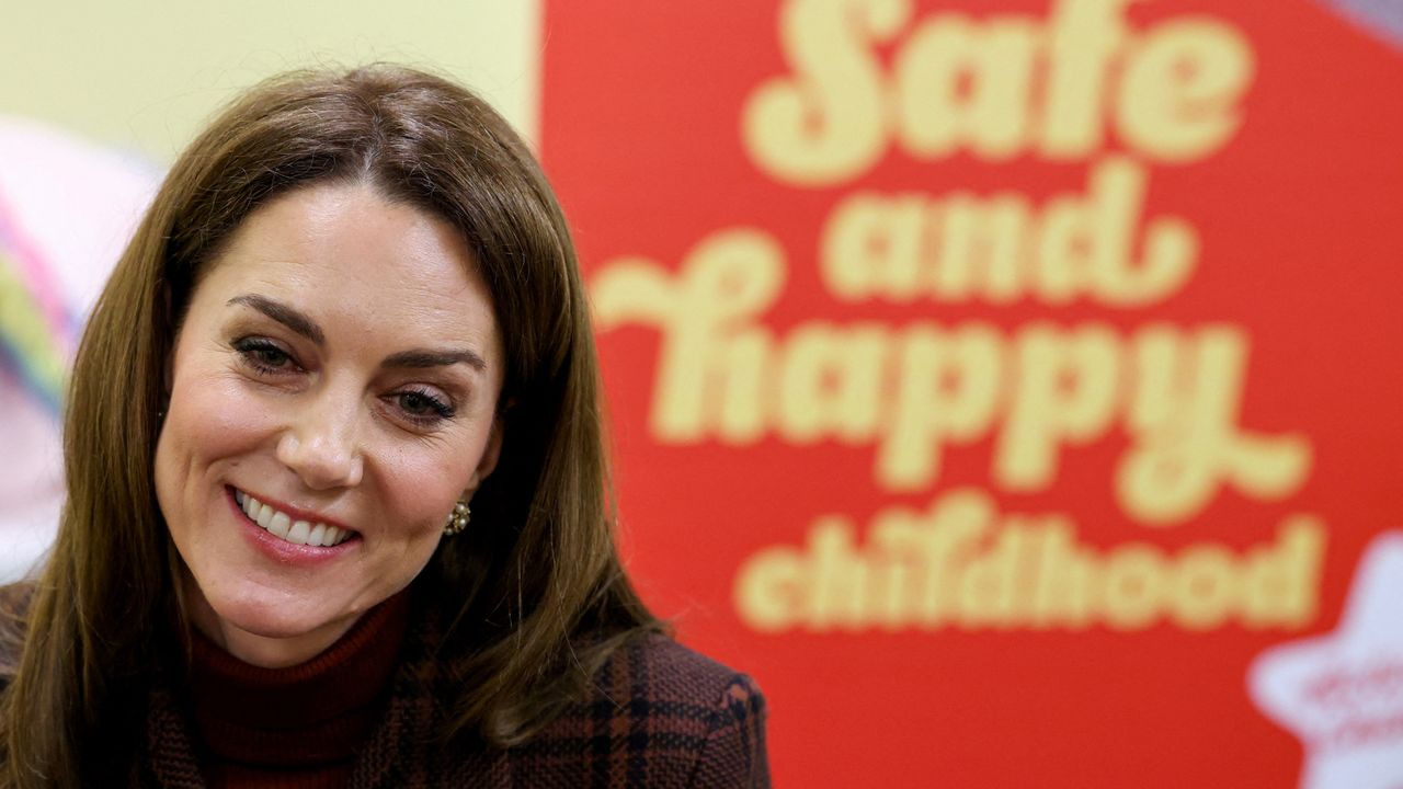 A shoulder up photo of Kate Middleton wearing a burgundy plaid coat sitting in front of a red sign saying safe and happy childhood in yellow letters