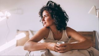 A woman with dark curly hair sitting up in bed with beige pyjama top and bedsheets with sun coming in through window