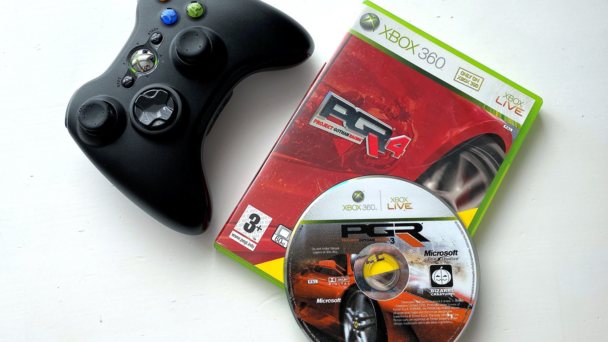 A black Xbox 360 controller next to the red cover of the video game "Project Gotham Racing 4" and a silver disc with a racing car design.
