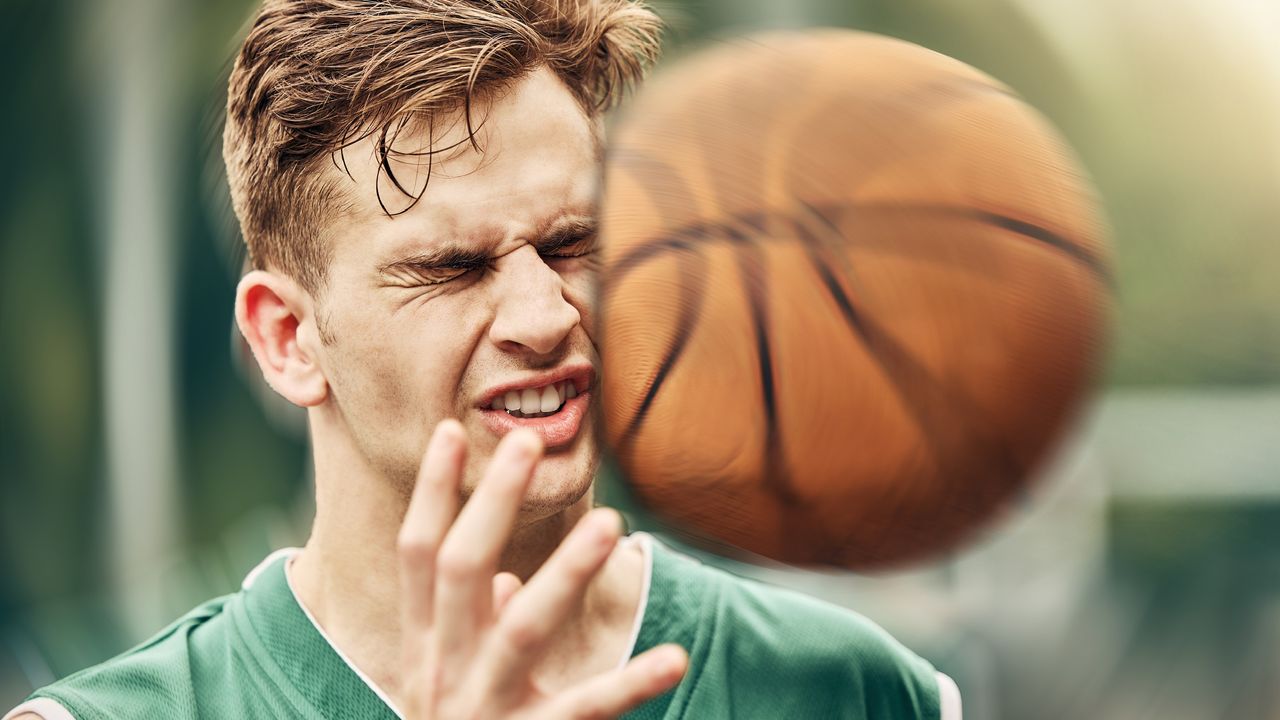 A man winces as he gets hit in the side of the head with a basketball.