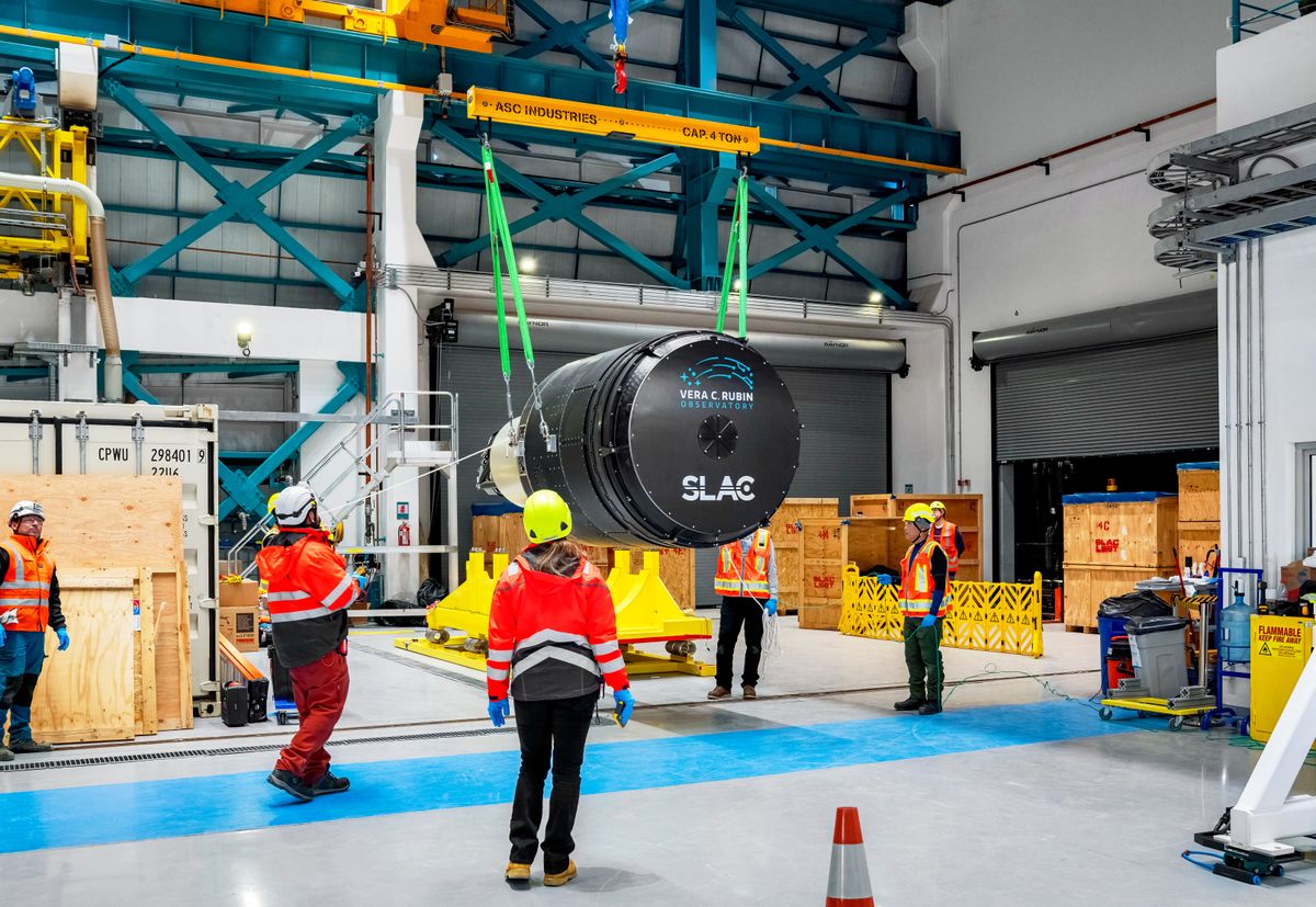 The car-sized LSST Camera is lifted above the floor inside Rubin Observatory, surrounded by about 8 people in construction protective clothing. The camera looks like a super-sized version of the lens portion of a handheld DSLR or similar, with a 5.5-foot diameter lens cap displaying the Rubin and SLAC National Lab logos.