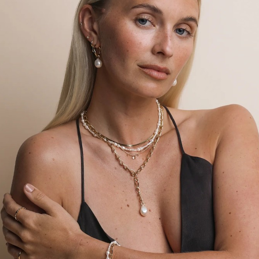A woman modelling Astrid & Miyu rings, bracelets, a necklace, and earrings while wearing a low-cut black slip dress 