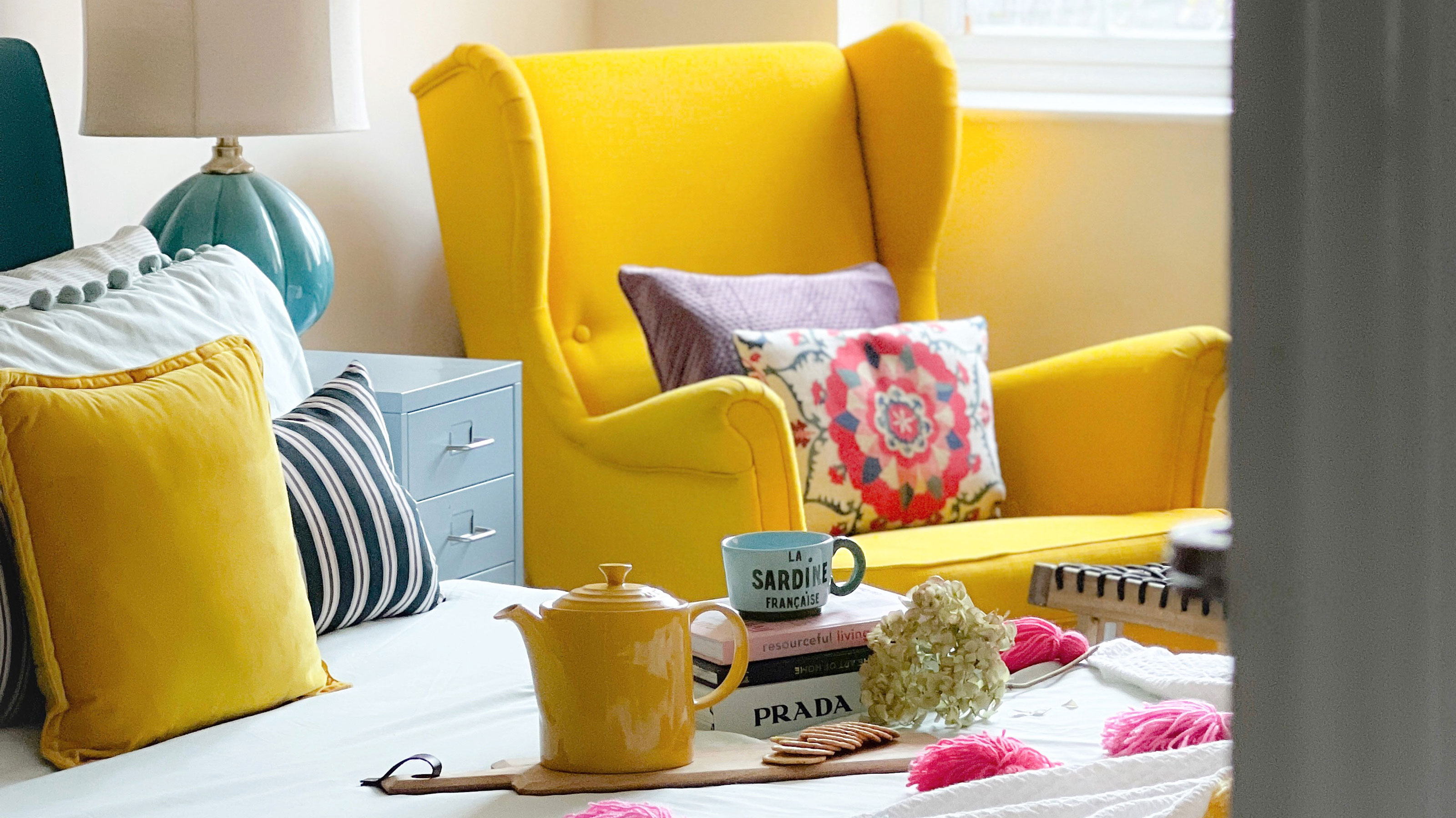 Yellow armchair behind bed with stack of books and yellow teapot resting on top