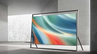 The 130-inch Samsung R95H Micro RGB TV displaying a colorful, abstract image in a gray-and-white, monochromatic room with no other objects. The TV is set within the large, easel-like frame that Samsung is selling with this TV.
