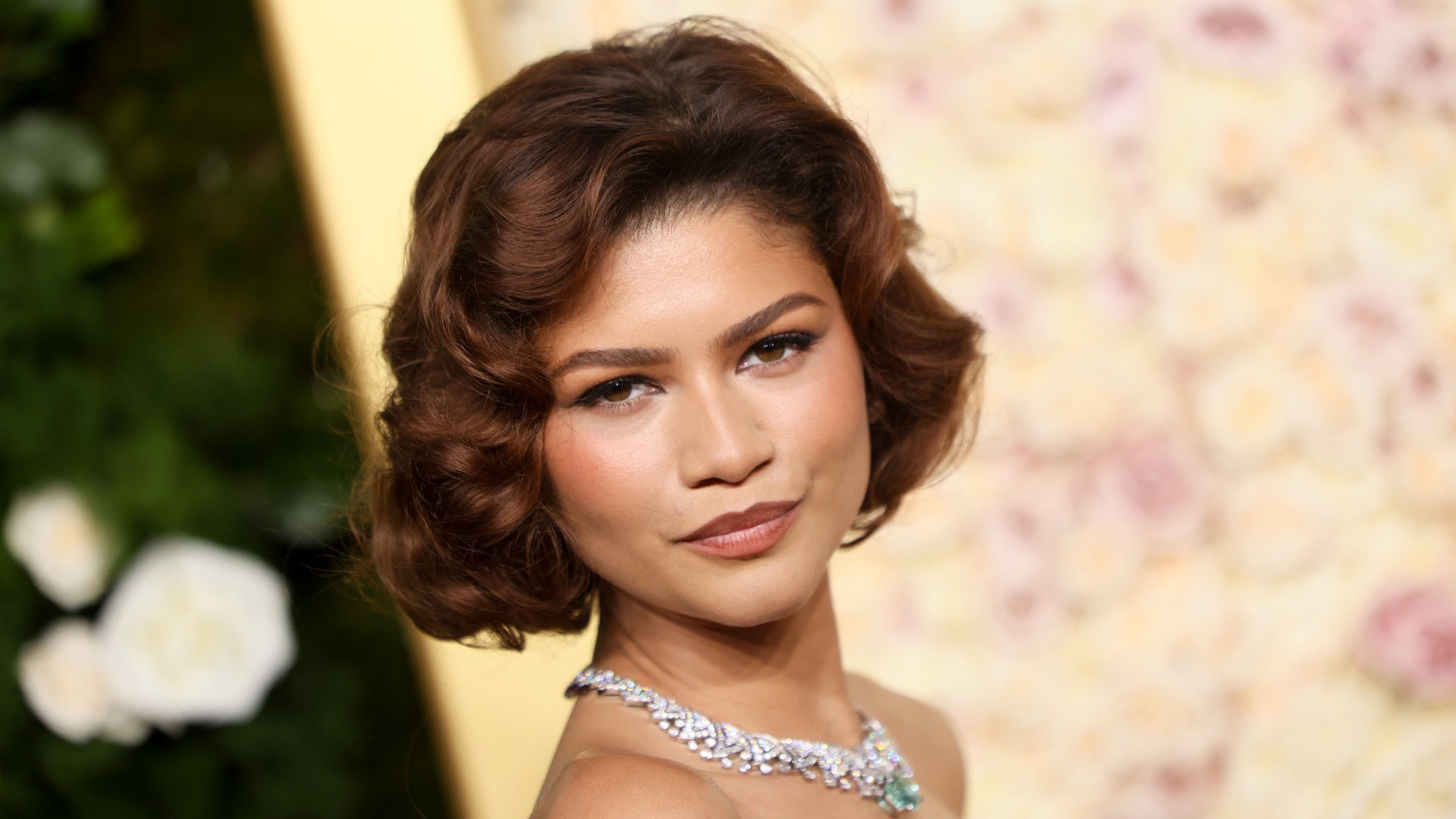 a close up of a light skinned woman with short hair posing in front of a floral background 