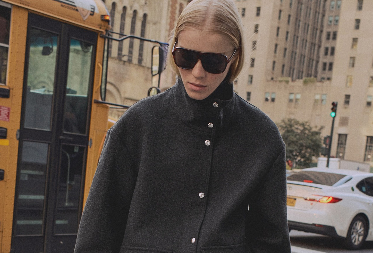 A Mango model wearing a gray funnel-neck jacket with silver buttons and large aviator sunglasses on the street in New York City.