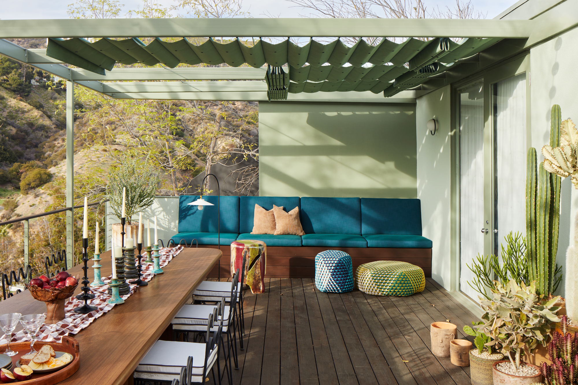 An avocado green outdoor space, with a covered seating area and large wooden dining table and multiple cacti with miss matched candle sticks.