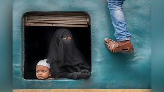 A woman wearing a burqa looking out of a train window with a child. A man's leg and foot is visible as he climbs on to the roof 