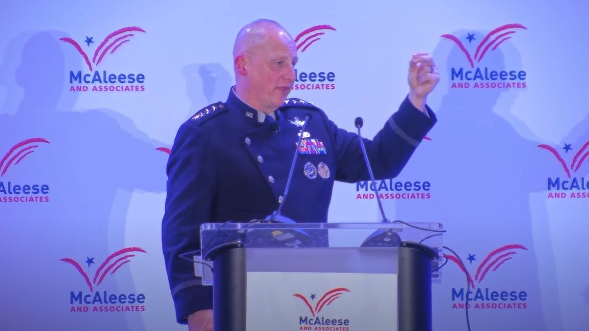 A man in military uniform holds up his bend arm behind a podium.