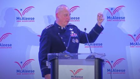 A man in uniform holds up his bend arm behind a podium.