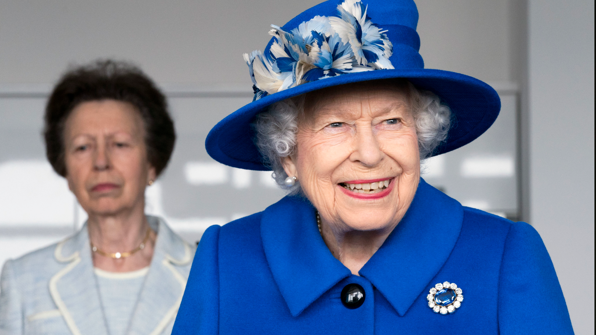 Queen Elizabeth smiling wearing a bright blue hat and coat with Princess Anne standing behind her grimacing