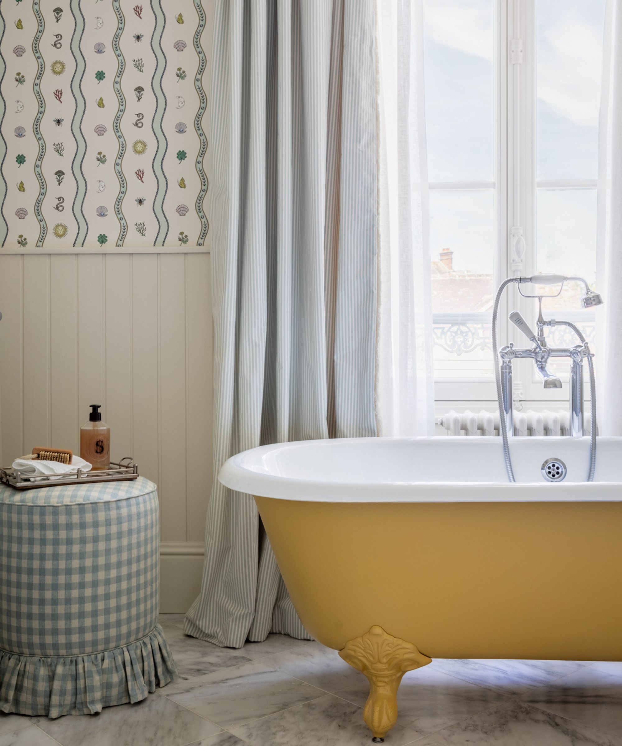 A light bathroom with a yellow clawfoot bathtub, marble floor, patterned wallpaper, and a blue gingham fabric-covered stool.