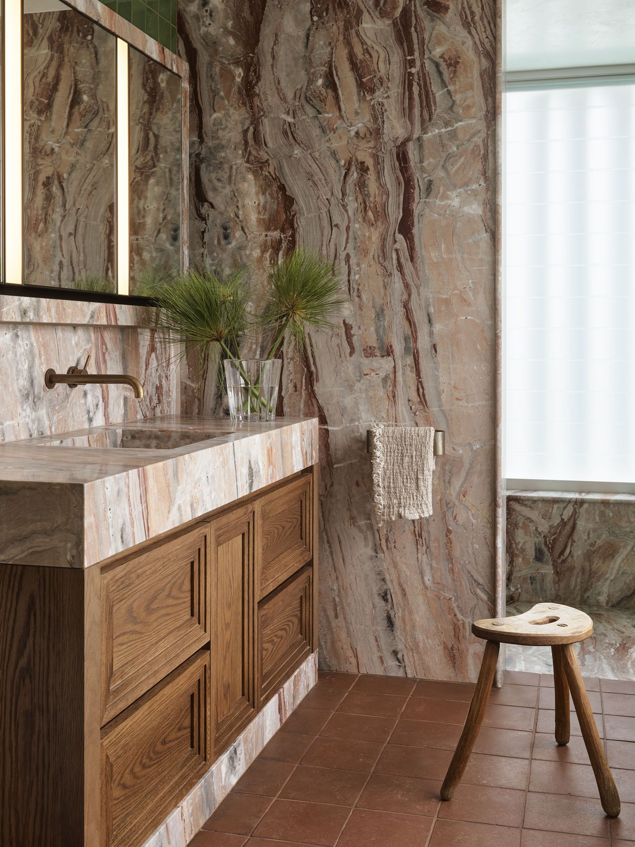 modern bathroom with marble wall and countertop, timber vanity, terracotta floor tiles, wooden stool, towel hook, vase with foliage, and mirror