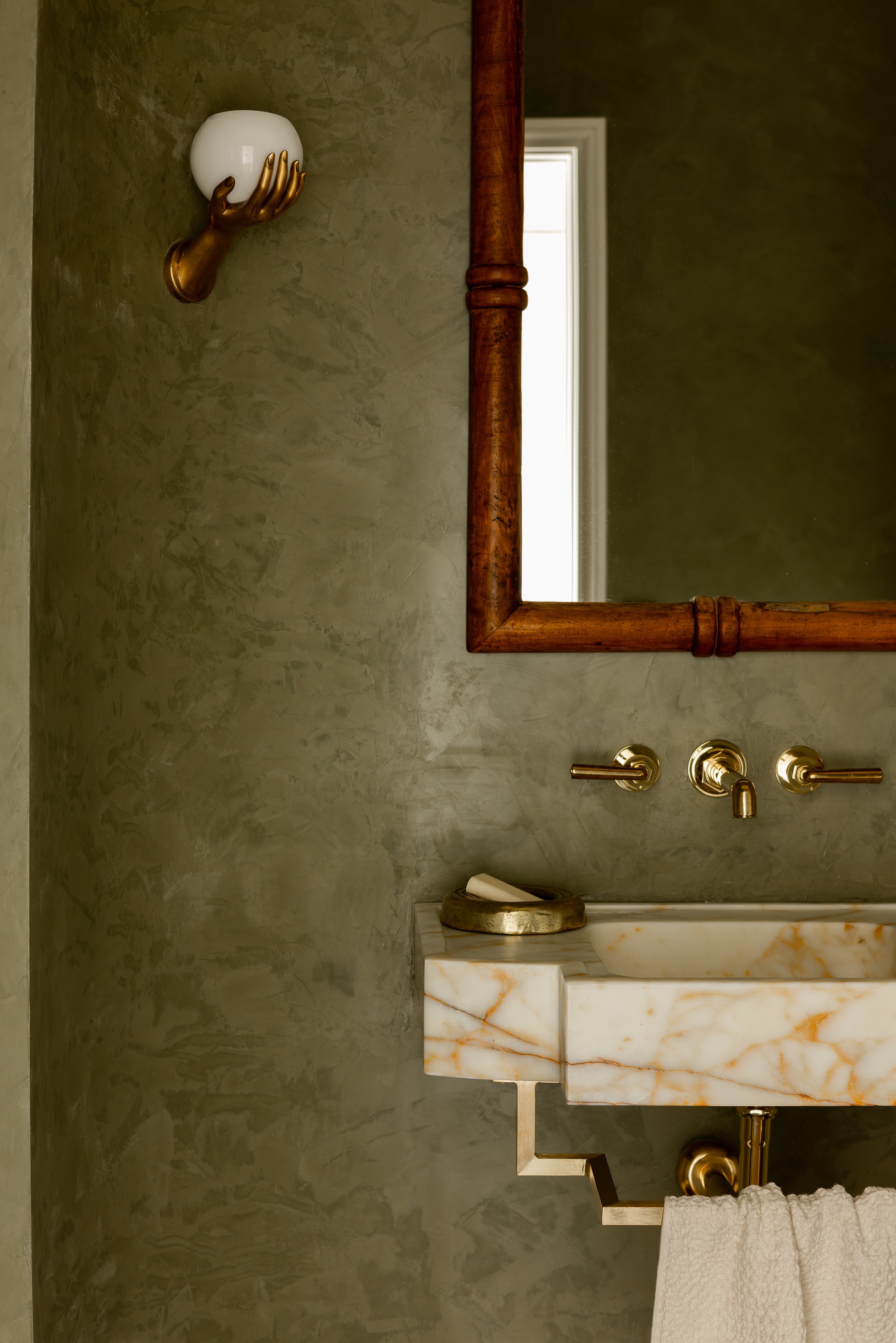 A green powder room with golden hardware, marble wall-mounted sink, wall sconce of hand holding light, a bamboo mirror