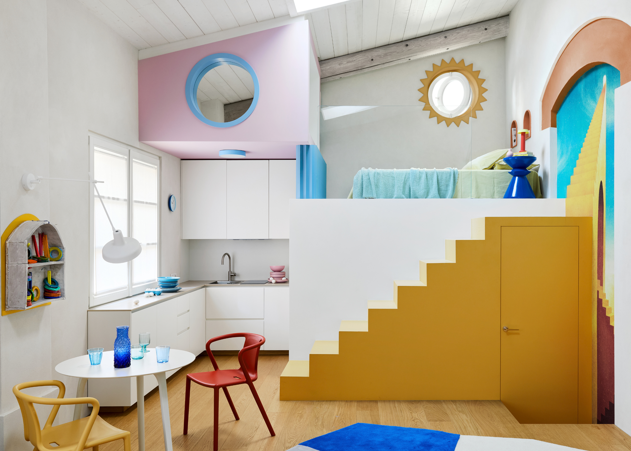 A white small apartment with a set of yellow stairs leading to a loft bed, a colorful mural on the wall, a small white dining table with colored chairs, and a white kitchen