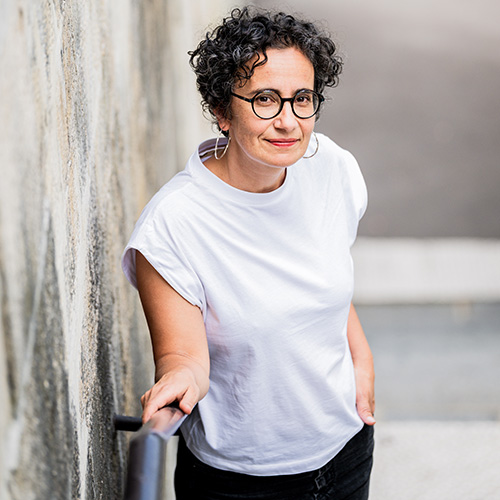 A woman with short curly hair and glasses wearing a white shirt looks at the camera