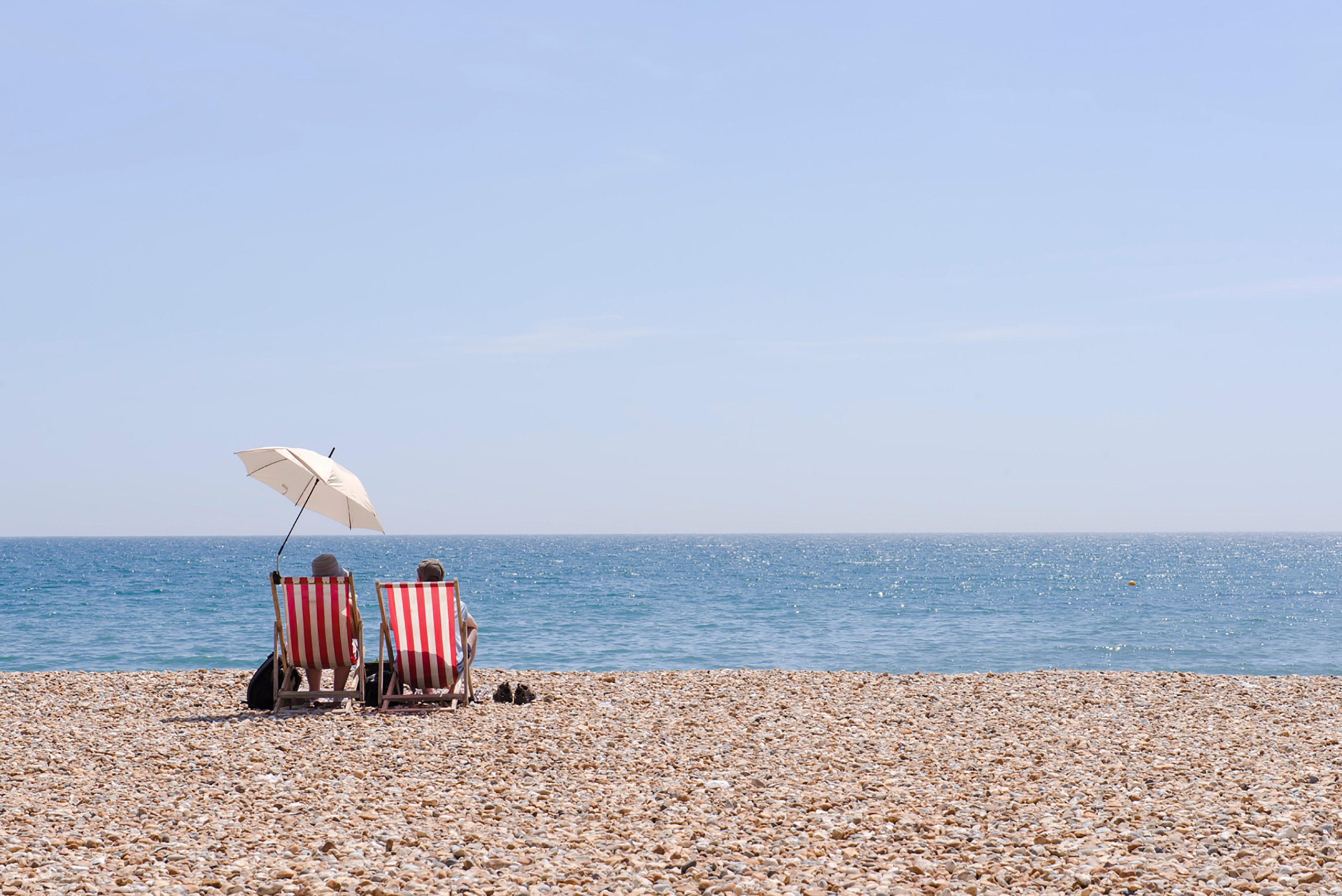 Two people on a shingle beach in the UK, staring out to sea under an umbrella