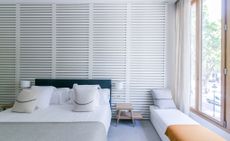 A hotel room with a bed, wooden side table, wall lamps, a white backless sofa, wooden wall slats and a large wood framed window.