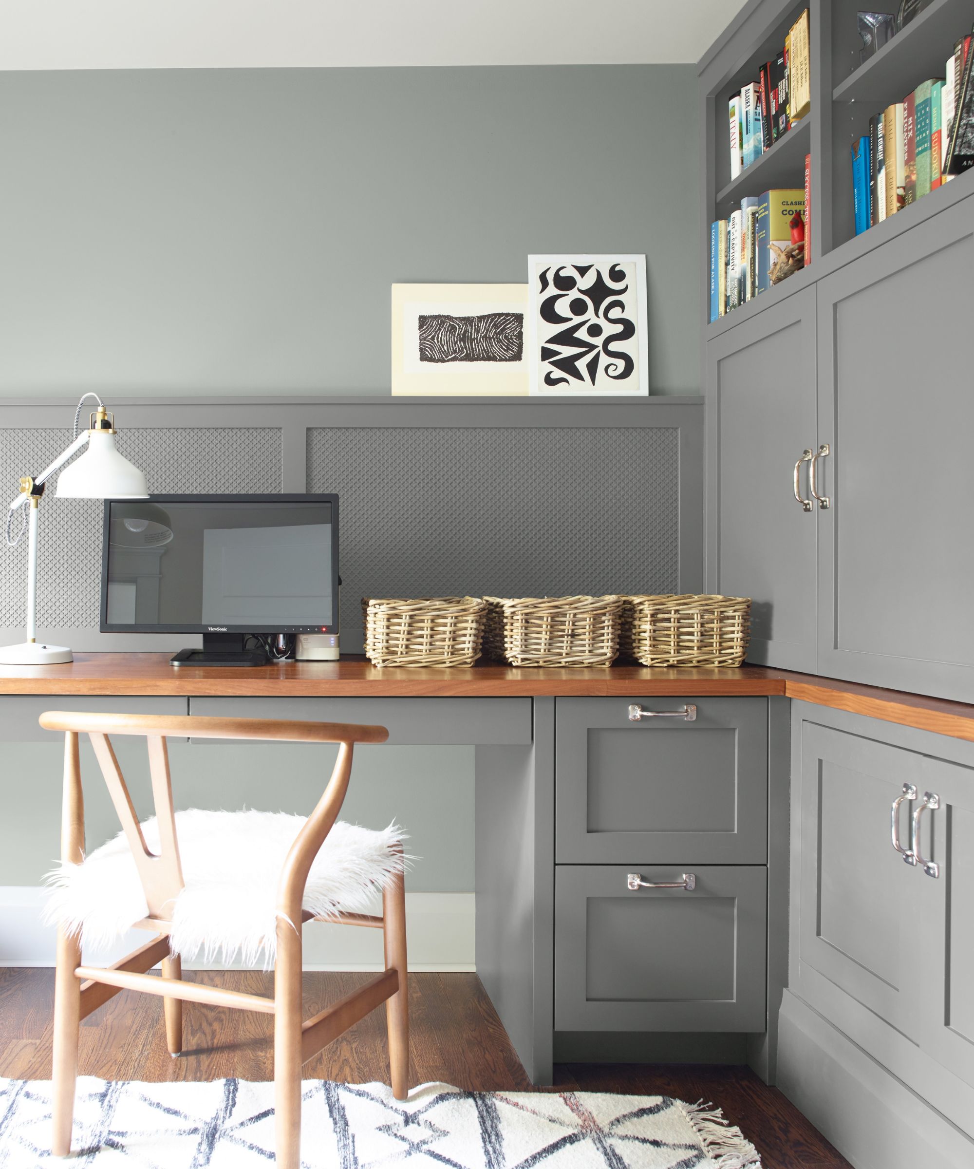 Stylish grey home office with built-in storage, wooden worktop, wicker baskets, bookshelves, and a timber chair with a fluffy white seat cushion.